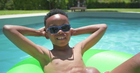 Boy relaxing on green pool float wearing sunglasses and red trunks on sunny day