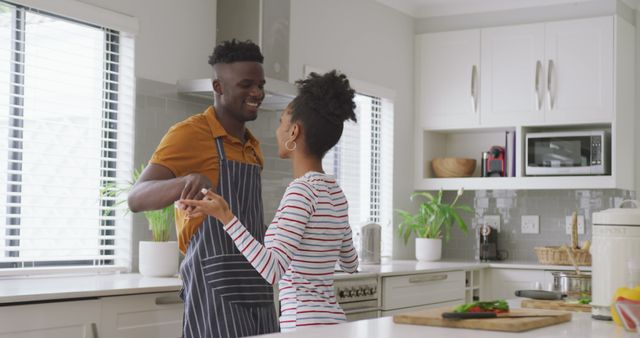 This photo depicts an African American couple smiling and dancing together in a modern, white kitchen. The scene exudes happiness and companionship, showcasing a great moment of leisure and intimacy in a domestic setting. This image can be used for advertisements related to home life, cooking, relationships, and lifestyle blogs emphasizing love, happiness, and joyful moments at home.