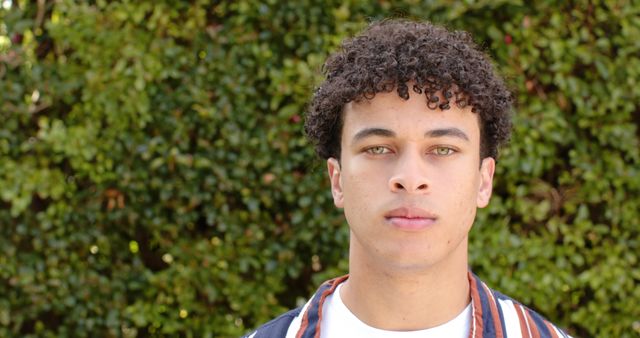 Close-up of a young man with curly hair standing in front of green foliage outdoors. The man has a serious expression, wearing casual clothing. This image can be used for concepts related to youth, confidence, determination, or nature backdrops. Suitable for promotional materials, blogs, websites, and social media posts focusing on lifestyle, fashion, or personal growth.