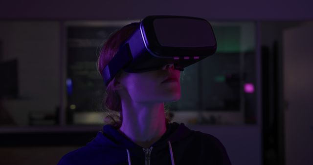 This image shows a woman using a VR headset in a dark room, experiencing a virtual reality environment. The dim lighting and shades of purple and pink give a futuristic and immersive feel to the scene. This visual can be used for showcasing VR technology, promoting tech products, illustrating articles on virtual reality and gaming, or for backgrounds in a tech presentation.