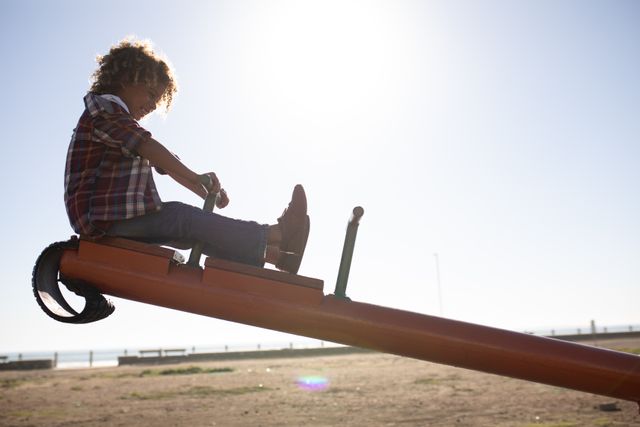 Side view of a biracial boy enjoying time playing at a playground, sitting on a see-saw on a sunny day by the sea