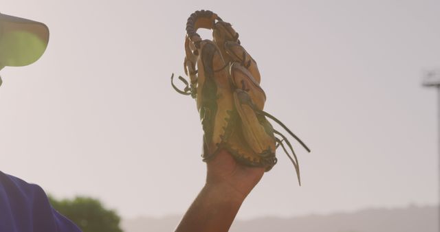 Photo portrays a baseball player's hand holding up a mitt against a clear sky, suggesting an outdoor sports setting in a sunny environment. Perfect for promoting sports-related activities, outdoor recreation, and summer games. Suitable for illustrating athletic equipment, baseball training, and competitive sports events.