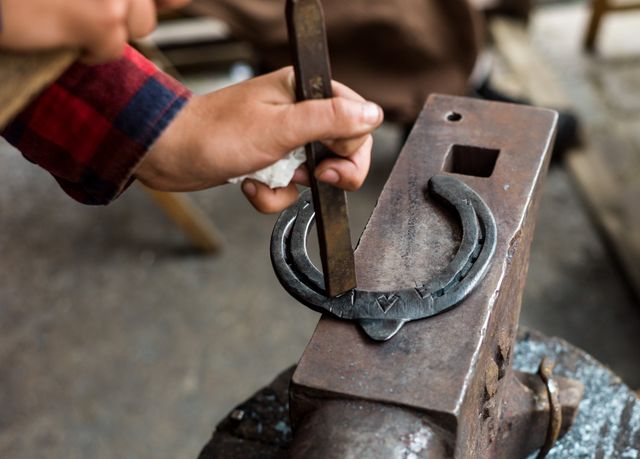 Image shows a blacksmith's hands skillfully crafting a metal horseshoe in a workshop. Ideal for use in articles or documentaries about traditional craftsmanship, skilled labor, blacksmithing techniques, or artisan trades. Great for illustrating themes of handmade work, metalworking, and historical trades.
