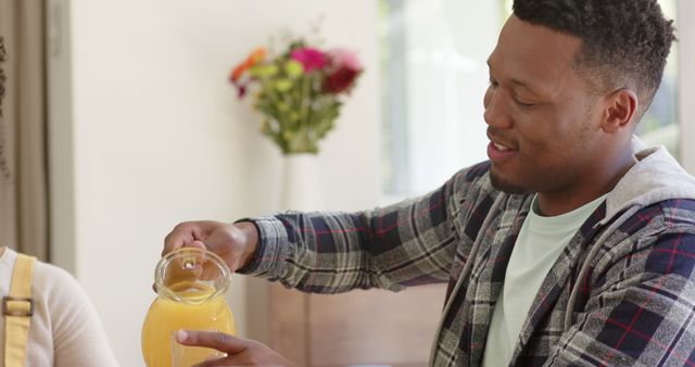Man pouring fresh orange juice into glass, likely at home during breakfast. Background includes a blurred bouquet of flowers on a wooden table. Ideal for depicting family meals, morning routines, and healthy lifestyle concepts.