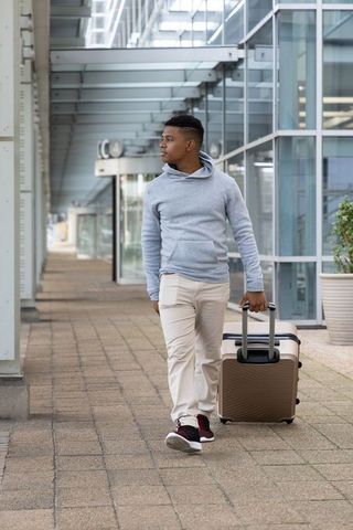 African american man walking with rolling suitcase along modern glass terminal corridor