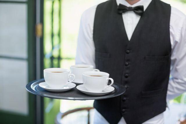Waiter in formal attire holding a tray with coffee cups in a restaurant. Ideal for illustrating themes of hospitality, professional service, dining experiences, and catering. Suitable for use in advertisements, brochures, and websites related to restaurants, cafes, and hospitality industry.