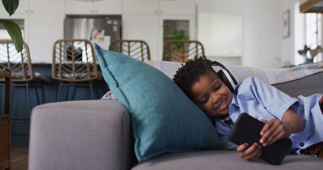 Happy child lying on a sofa, relaxing with a smartphone and wearing headphones. Perfect for illustrating modern technology use among kids, home leisure activities, or youthful relaxation settings.