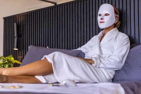 Woman relaxing on bed wearing led facial mask and white robe for skincare and relaxation