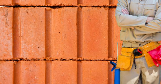 Construction worker with arms crossed standing confidently in front of brick wall background. Ideal for use in construction company promotions, labor services advertisements, training materials, and handyman service posters. Emphasizes professional skills, building work, and craftsmanship.