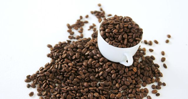 Freshly roasted coffee beans overflowing from a white cup onto a white background, creating a striking contrast. Ideal for use in coffee shop promotions, breakfast menu designs, blog posts on coffee culture, or any marketing materials related to beverages and morning routines.