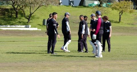 Diverse male cricket teammates huddling on grassy oval wearing black jerseys and batting pads