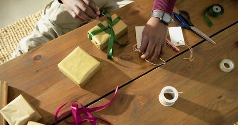 Mid-adult African American man wrapping gold-wrapped gifts with green ribbon on wood table