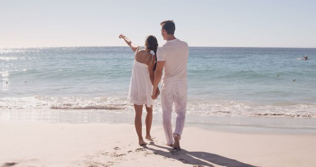 Couple in casual attire holding hands and walking along sandy beach. The woman points towards horizon while the man walks beside her. They are enjoying a sunny summer day at the sea. Ideal for content related to romance, vacations, travel destinations, relationship goals, beach activities, or promoting summer getaways.