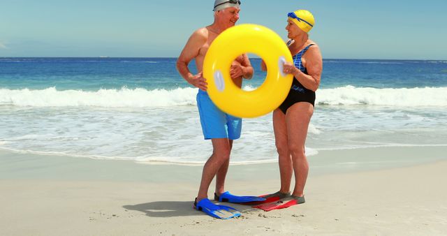 Senior couple standing on sandy beach wearing swim gear, holding yellow inflatable ring, and smiling. Perfect for promoting active lifestyle, travel destinations, senior fitness, and summer vacation activities.
