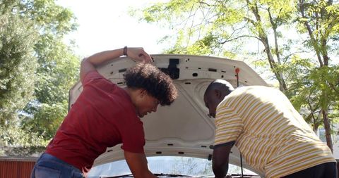 Two men inspecting car engine in driveway repairing sedan under sunny trees
