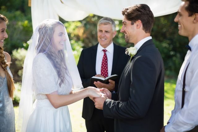 Bride and groom exchanging vows, holding hands, outdoors with minister. Ideal for wedding invitations, celebratory announcement designs, event planning materials, and romance-themed promotions.