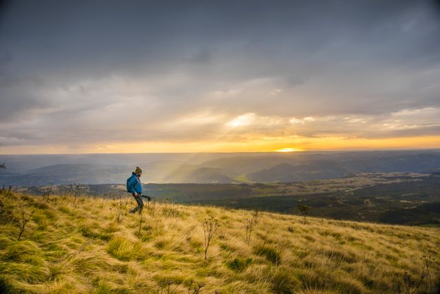 Hiker overlooking a scenic sunset from a grassy hill. Puffy clouds cast shadows as sunlight breaks through, creating a golden glow on the landscape. Excellent for themes of adventure, tranquility, and natural beauty. Can be used in environmental campaigns, travel promotions, or outdoor lifestyle blogs. Perfect for inspiring wanderlust and appreciating natural landscapes.