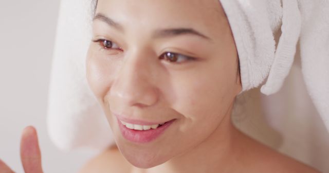Image of portrait of smiling biracial woman with towel on hair applying face serum in bathroom. Health and beauty, leisure time, domestic life and lifestyle concept.