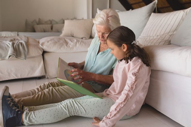 Grandmother and granddaughter sitting on floor, reading storybook together in cozy living room. Ideal for illustrating family bonding, intergenerational relationships, and educational activities at home. Perfect for use in family-oriented content, educational materials, and lifestyle blogs.