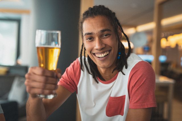 Young biracial man with dreadlocks smiling and holding a glass of beer in a restaurant. Ideal for use in advertisements for social gatherings, restaurants, bars, or lifestyle blogs focusing on leisure and friendship.
