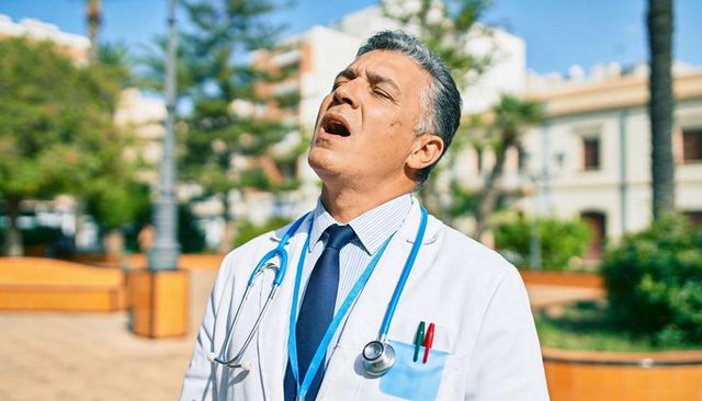 Senior doctor taking deep breath outdoors in sunlit plaza wearing stethoscope