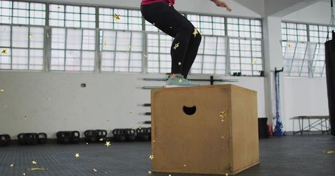 Athlete performing box jump on wooden plyo box in gym wearing black leggings and teal-gray shoes