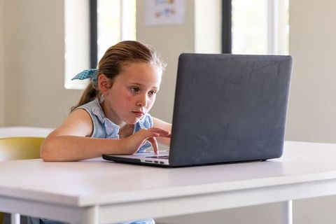 Young girl studying on laptop at white desk for remote learning and homeschooling