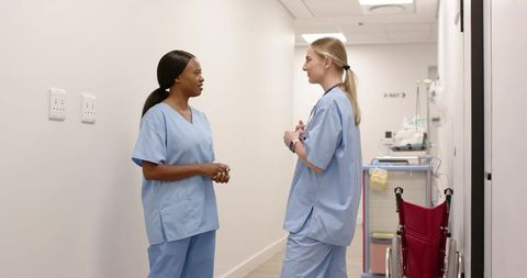 Two nurses discussing patient care in hospital corridor with wheelchair and medical cart