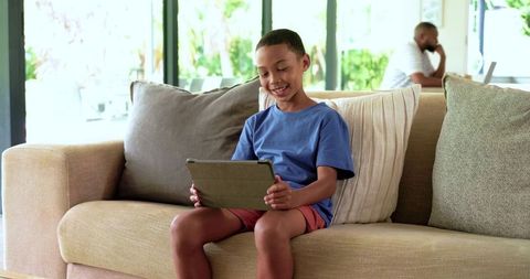 African american father working from home while son relaxing on sofa with tablet