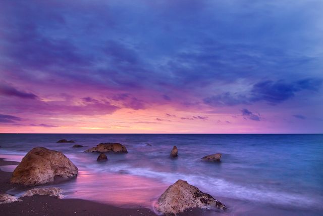 This colorful scene captures the calm waves of the ocean gently washing over rocks on a beach at sunset. The sky is a vibrant blend of purples, pinks, and oranges, creating a serene and tranquil atmosphere. Useful for backgrounds, travel promotions, relaxation themes, and nature-based projects.