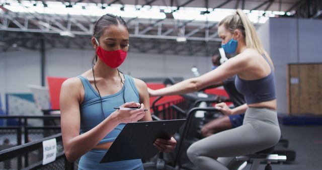 Women in athletic clothing are working out at a gym while wearing face masks. One woman is writing on a notepad, possibly tracking progress or training plans, while others are using cardio equipment like treadmills. This image is perfect for usage in fitness-related content, health and safety guidance on working out with precautions, or articles promoting active lifestyles during health-conscious times.