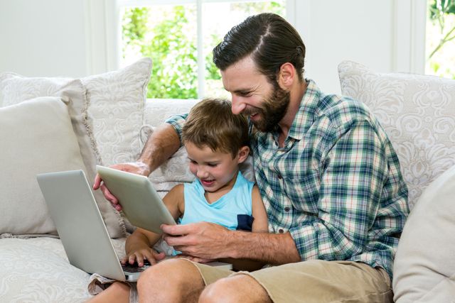 Playful son and father with laptop and digital tablet on sofa - Download Free Stock Photos Pikwizard.com