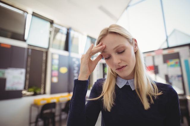 Blonde female teacher standing in classroom, looking stressed and thoughtful. Ideal for use in articles or advertisements related to education, teacher stress, mental health in the workplace, and stress management strategies for educators.