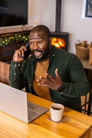 African american man working remotely, talking on phone and gesturing at laptop in cozy home office