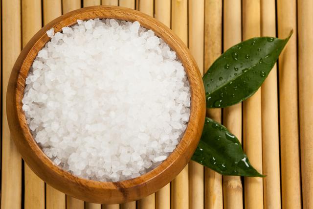 Sea salt in a wooden bowl placed on a bamboo mat with green leaves. This image is ideal for use in wellness, spa, and self-care content. Perfect for promoting natural and organic products, relaxation techniques, and detox treatments.
