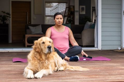 Mature woman practicing calm outdoor yoga on porch with golden retriever and smartphone