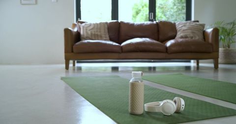 Beige reusable water bottle and white headphones resting on green yoga mat in living room