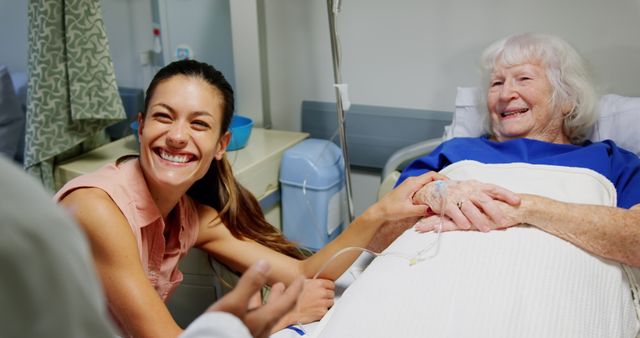 Image depicts a joyful caregiver holding the hand of an elderly woman in a hospital bed. Ideal for use in healthcare promotion, elderly care awareness, hospital marketing materials, and brochures on compassionate care and patient well-being.