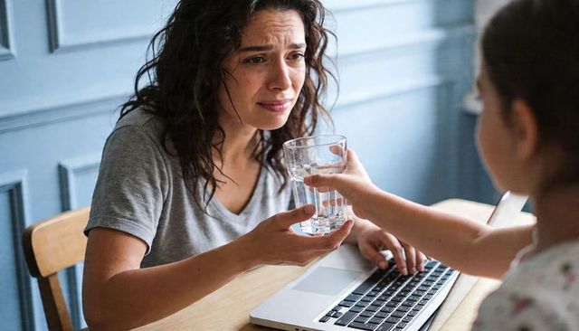 Overwhelmed woman accepting glass of water from child while working on laptop at home
