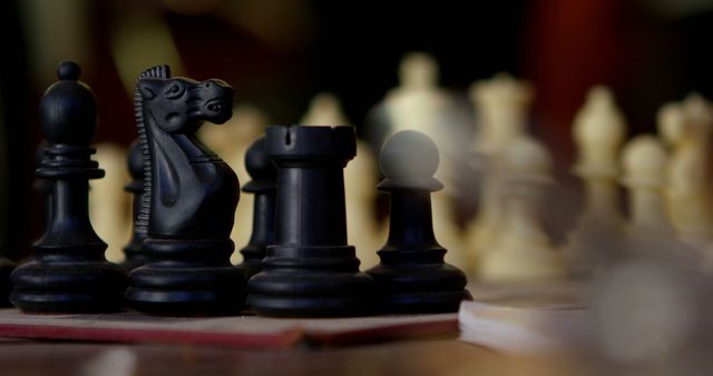 Close-up view of chess pieces on wooden board, focusing on black pieces: knight, pawn, and rook. The image has a soft focus background that includes blurred white pieces. This can be used for content related to strategy, games, intellectual pursuits, competitions, and thinking skills.