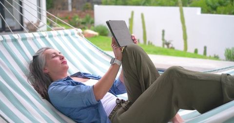 Mature woman relaxing in striped hammock reading tablet on sunlit backyard patio