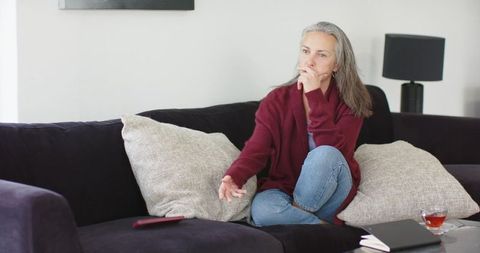 Thoughtful mature woman relaxing on sofa reaching for remote in cozy living room