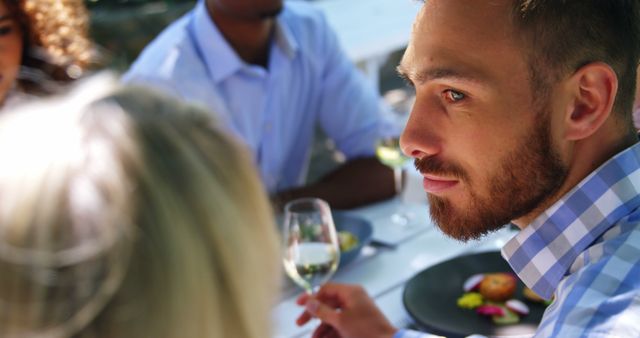 Man engaging in conversation with friends outdoors, enjoying a meal and a glass of wine. Ideal for uses related to social interactions, outdoor dining, summer gatherings, and lifestyle promotions.