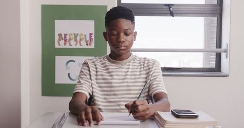 Focused African American Teen Writing in Classroom with Books and Smartphone