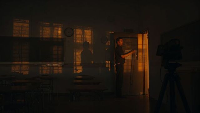 Standing teacher watching golden window light in empty classroom at dusk, cinematic mood