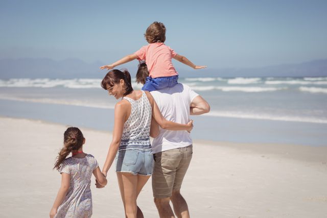 Family enjoying a sunny day at the beach, walking along the shore. Ideal for use in travel brochures, family vacation advertisements, and lifestyle blogs promoting outdoor activities and family bonding.