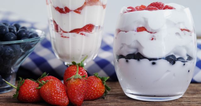 Two glasses of layered yogurt parfaits with fresh strawberries, blueberries, and strawberry sauce, with copy space. A checkered blue and white napkin adds a cozy, casual touch to this delicious and healthy snack setup.