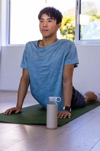 Asian man performing prone backbend on yoga mat at home wearing smartwatch with bottle