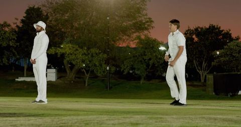 Two cricketers standing on dusk turf under floodlights wearing white kit and bucket hat