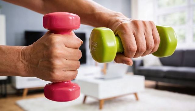 Middle-aged hands holding colorful dumbbells during bright living room home workout
